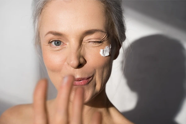 Uśmiechnięta kobieta z widocznymi zmarszczkami wokół oczu pokazuje, jak aplikuje na twarz krem z peptydami